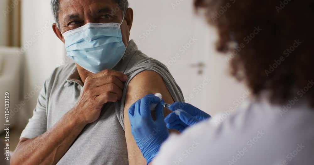Mixed race female doctor wearing mask giving vaccination to senior man at home