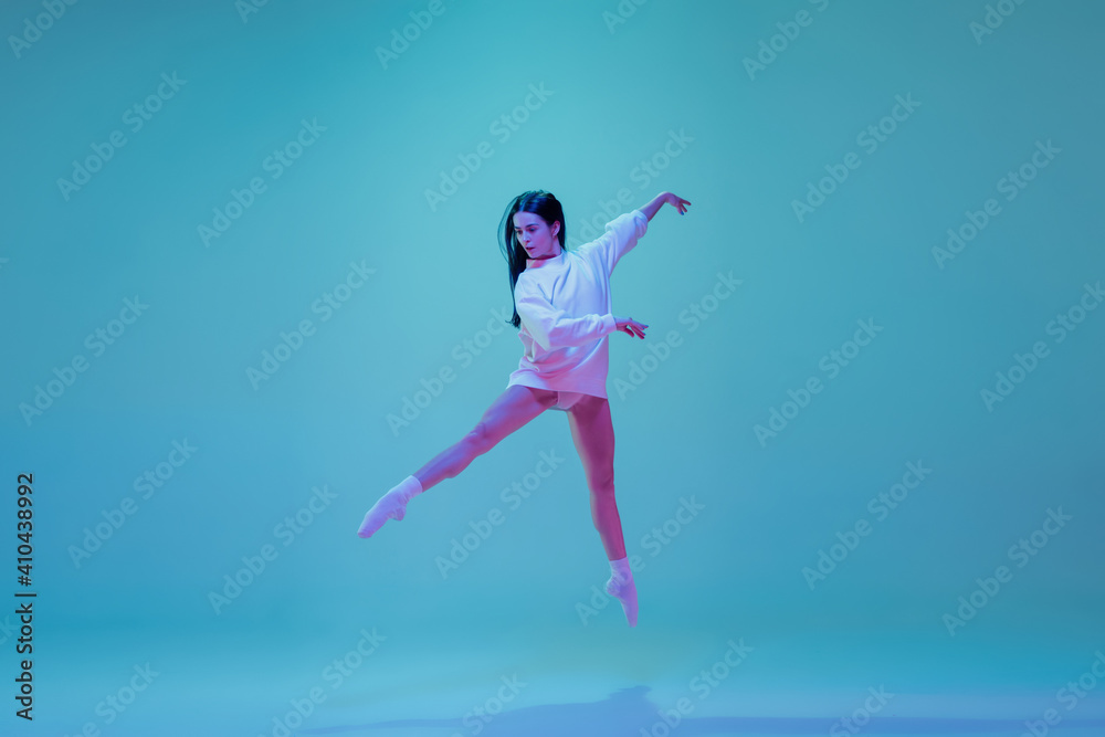 Obraz premium In flight. Young and graceful ballet dancer isolated on blue studio background in neon light. Art, motion, action, flexibility, inspiration concept. Flexible caucasian ballet dancer, moves in glow.