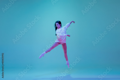 In flight. Young and graceful ballet dancer isolated on blue studio background in neon light. Art, motion, action, flexibility, inspiration concept. Flexible caucasian ballet dancer, moves in glow.