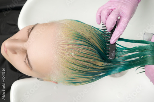 Top view of hairstylist holds wet hair in hand, combs long green and discolored hair of client while washing hair in shower in special sink. Working professional hairdresser in pink protective gloves.