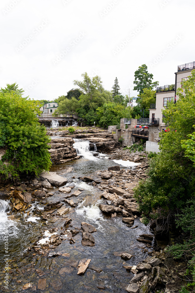 wide angle view of Almonte Falls, Ontario, Canada, a tiered and multiple segmented waterfall, surrounding small trees and grasses on bedrock during summer.
