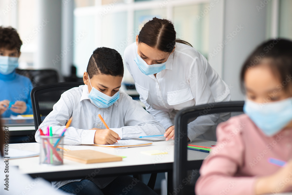 Female teacher in disposable face mask helping small boy Female teacher in disposable face mask helping small boy