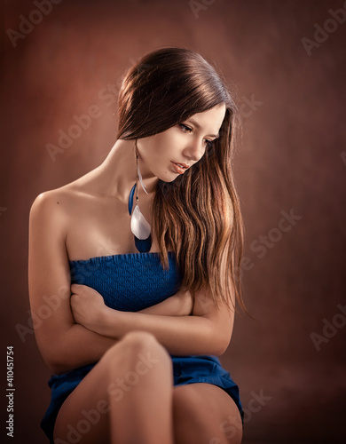 a girl with a jewelry in her ear in a blue dress sits and looks down on a brown background