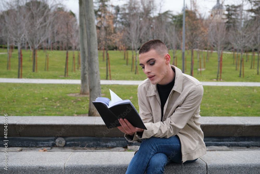 Handsome young man wearing make up, reading a book sitting on a wall. Non binary androgynous guy. Handsome young man wearing make up, reading a book sitting on a wall. Non binary androgynous guy.