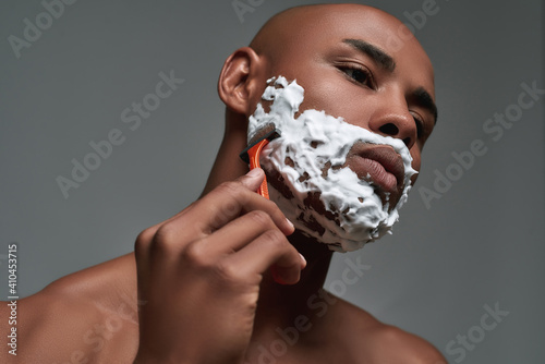 Close up portrait of handsome young african american man with foam applied on his face using steel razor for shaving isolated over gray background
