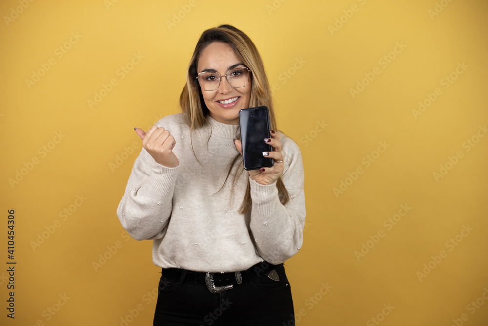 Obraz premium Pretty woman with long hair holding smartphone showing blank screen smiling happy pointing with hand and finger to the side against yellow wall