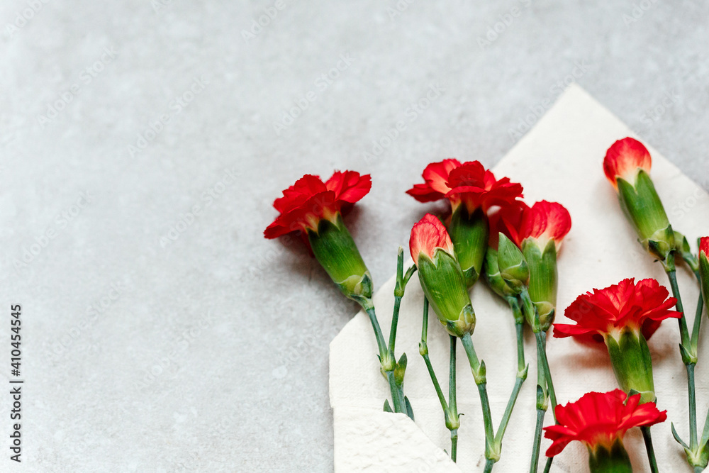 Rote Nelken Blüten in einem cremefarbenen Briefumschlag aus strukturiertem Naturpapier - von oben auf grauem Untergrund Rote Nelken Blüten in einem cremefarbenen Briefumschlag aus strukturiertem Naturpapier - von oben auf grauem Untergrund