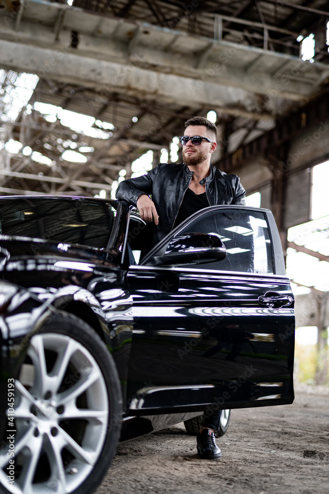 Portrait of a stylish bearded man in trendy sunglasses and leather jacket stands near black car. Fashion portrait on abandoned warehouse background.