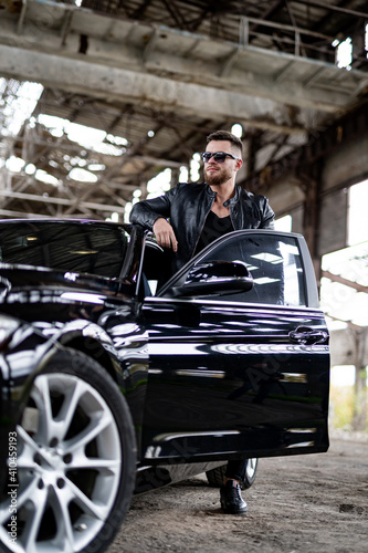 Portrait of a stylish bearded man in trendy sunglasses and leather jacket stands near black car. Fashion portrait on abandoned warehouse background. © Vadim