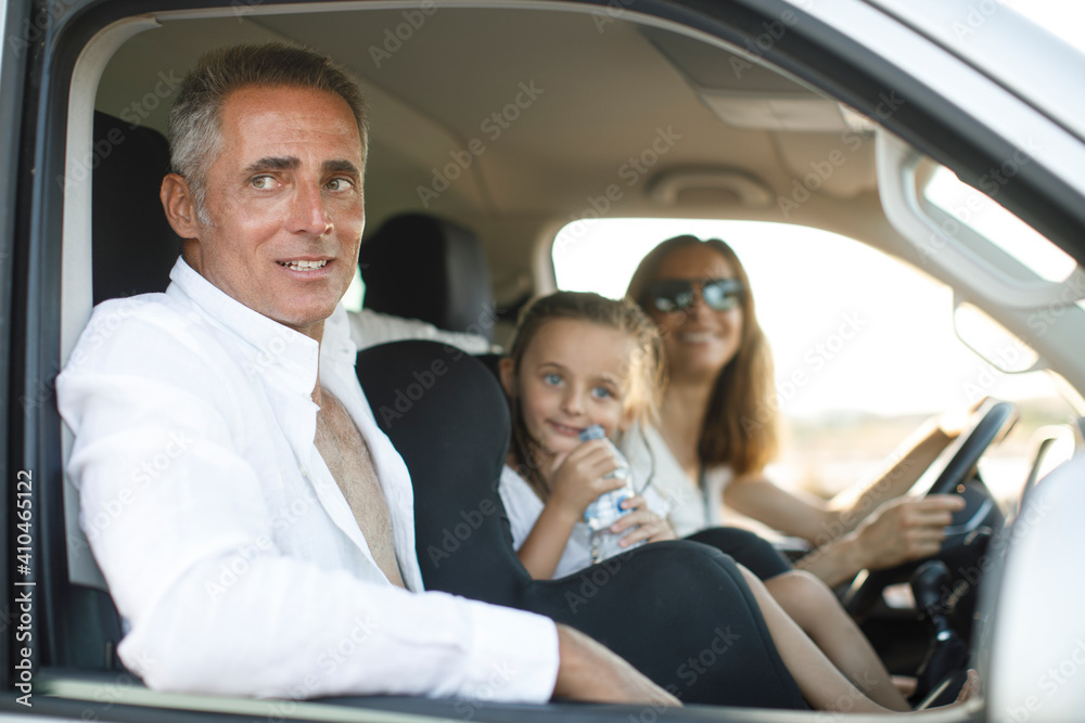 Donna al volante con maglietta bianca in contesto vacanziero viaggia con la figlia e il marito con espressioni sorridenti seduti all'interno dell'abitacolo di un furgone