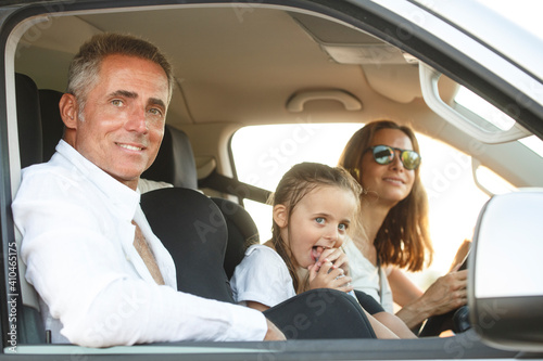 Donna al volante con maglietta bianca in contesto vacanziero viaggia con la figlia e il marito con espressioni sorridenti seduti all'interno dell'abitacolo di un furgone