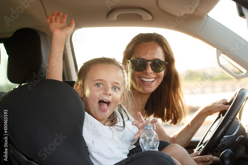 Donna al volante con maglietta bianca in contesto vacanziero viaggia con la figlia con espressioni sorridenti salutando qualcuno in lontananza photo