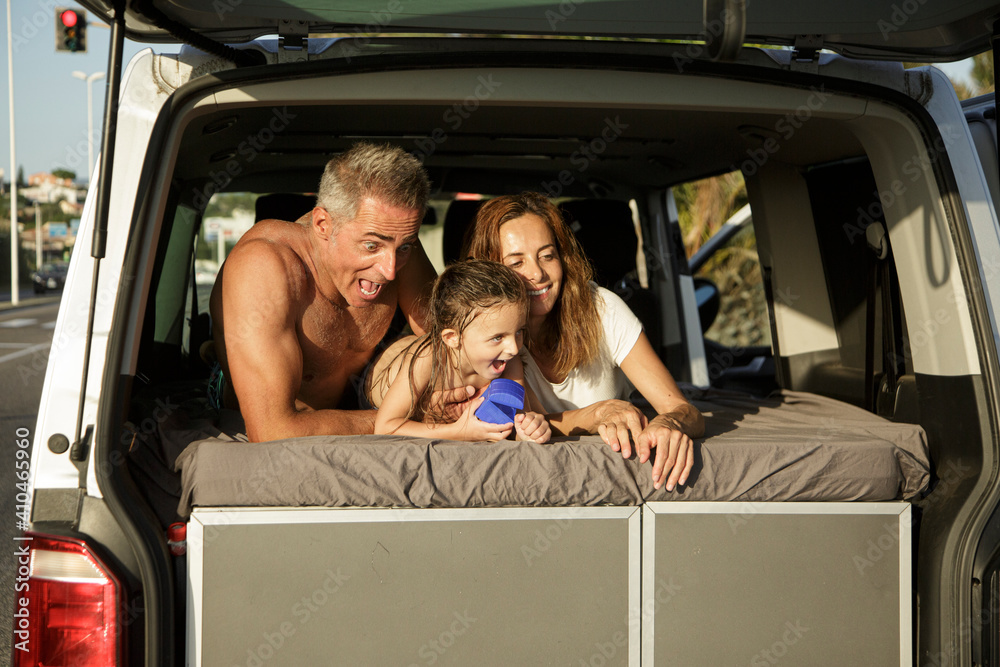 famiglia felice composta da papà mamma e figlia, in abbigliamento da mare si abbraccia felice dentro al furgone parcheggiato in strada