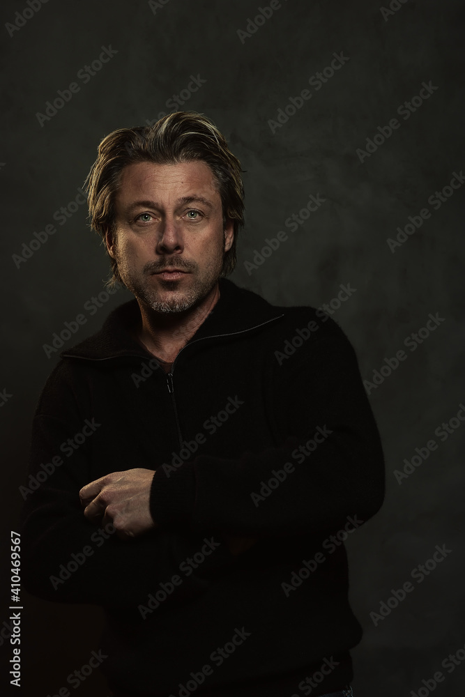 Blonde man in black woolen sweater in front of dark grey wall.