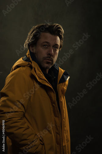 Shadowy portrait of blonde man in yellow coat in front of grey wall.