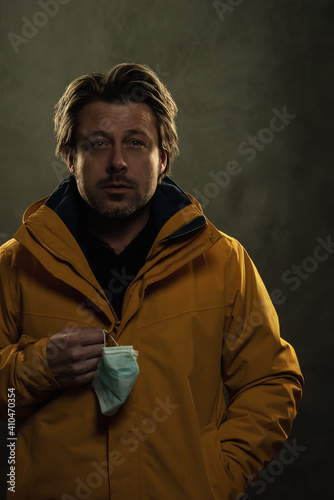 Shadowy portrait of blonde man holding hygienic face mask wearing a yellow coat in front of grey wall.