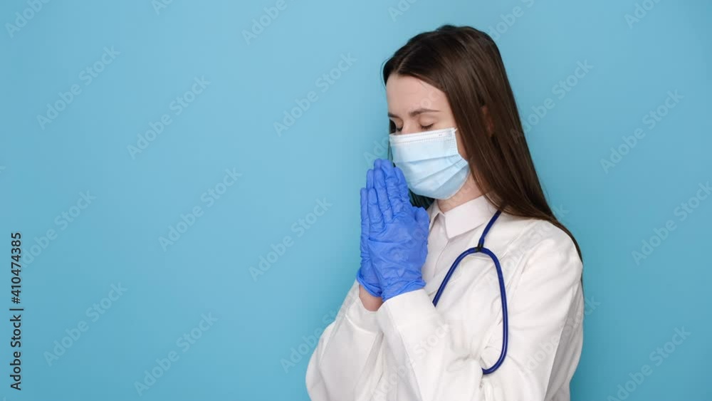 Female doctor in white medical gown sterile face mask gloves holding hands folded in prayer, isolated over blue background. Epidemic pandemic coronavirus 2019-ncov sars covid-19 flu virus concept Female doctor in white medical gown sterile face mask gloves holding hands folded in prayer, isolated over blue background. Epidemic pandemic coronavirus 2019-ncov sars covid-19 flu virus concept