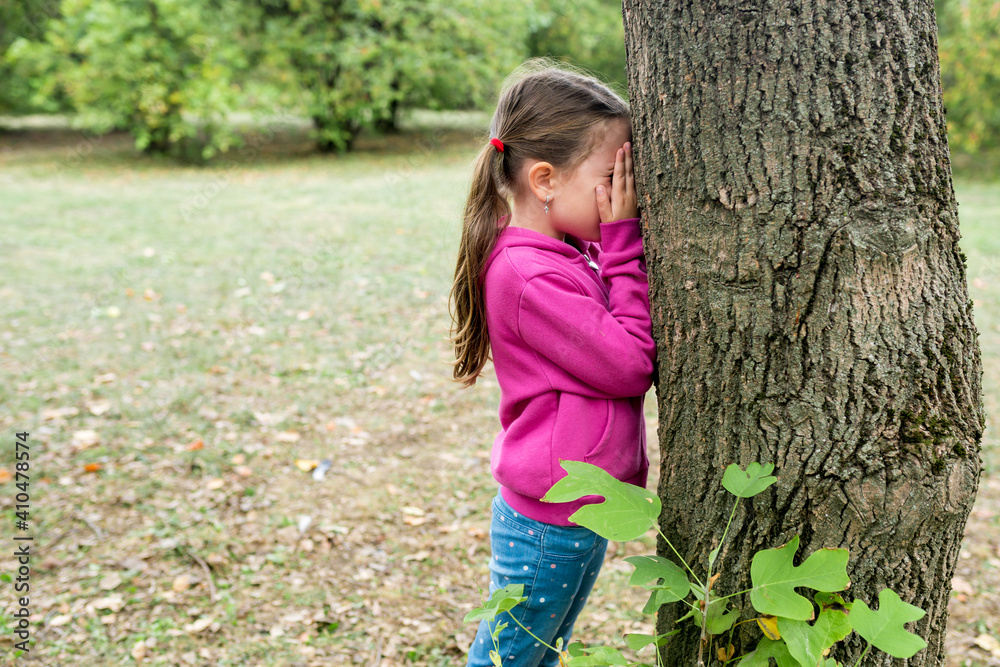 Children playing hide and seek in the park on an autumn day. The girl standing near a tree and covering her face with hands. Outdoor activities with kids Children playing hide and seek in the park on an autumn day. The girl standing near a tree and covering her face with hands. Outdoor activities with kids