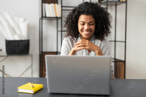 Smiling African American female freelancer or student with Afro hairstyle sitting at the desk in the home office, studying, taking an online course, listening to the webinar, looking at laptop screen