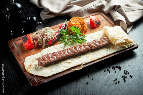 Grilled Turkish lamb lula kebab with grilled vegetables, onion and rice on a wooden board. Dark background.