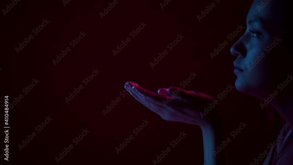 Pretty female blowing magic sparkling colourful glitter from palms in neon studio light, close up with copy space