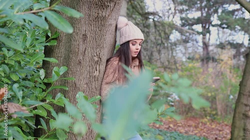 A woman on her phone in the winter time wearing a woolly jumper and pink bobble hat in a wooded park with somebody looking and spying at her through branches and bushes, voyeurism stalking concept.