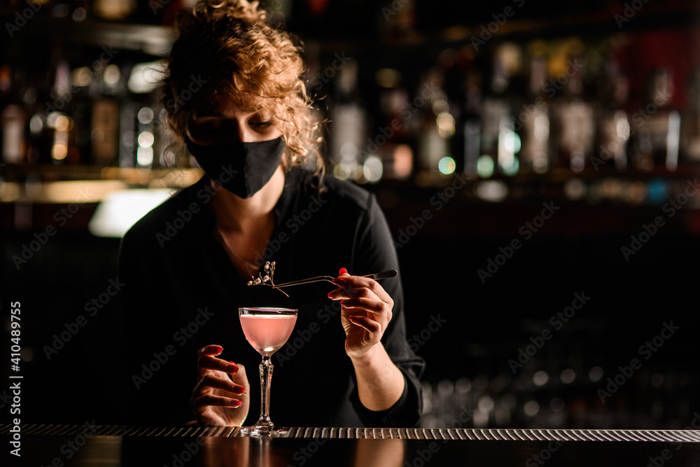 view of woman bartender in mask that neatly decorates glass of cocktail with flower sprig