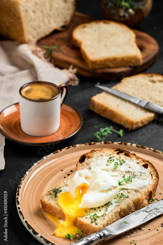 Soft-boiled (poached) egg on slice of bread covered with butter cream and herbs, on clay plate on black wooden table. Espresso coffee and loaf of sliced bread on blurred background. Breakfast idea