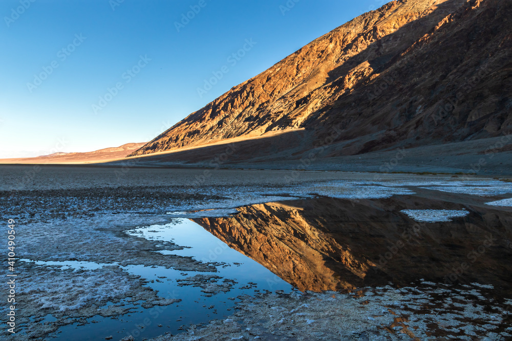 Badwater Basin, Death Valley National Park. Crystal deposits ring open water mirroring the hills in the distance. Blue sky above. 
