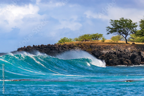 Wave breaking on Kona coast of Hawaii's Big Island. Trees on rocky ledge behind. Cloudy sky in the background. 