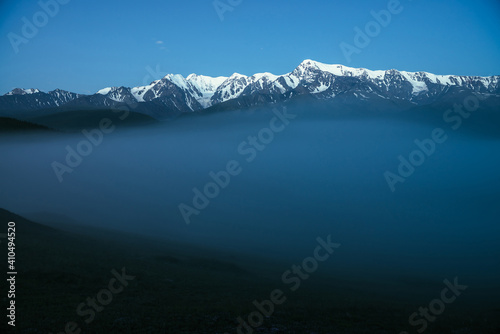 Atmospheric mountains landscape with dense fog and great snow mountain top under twilight sky. Alpine scenery with big snowy mountains over thick fog in night. High snow pinnacle above clouds in dusk.