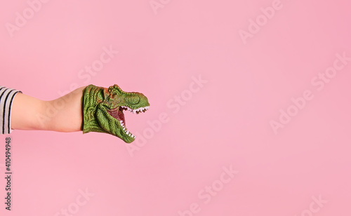 Fototapeta Naklejka Na Ścianę i Meble -  Kid hand wearing funny dinosaur puppet doll on pink background. Dinosaur toy growls roar and speaks. Copy space for advertisements and text promotional content.