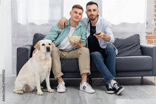 cheerful same sex couple watching movie with popcorn near labrador