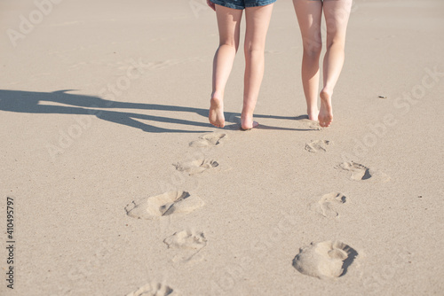 Image of female feet close-up on a beach background. Two women walk barefoot on the sand, leaving footprints.