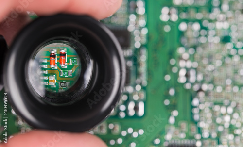 Small electronic components in black loupe on green PCB background. Closeup of magnified red semiconductor diodes, resistor or capacitors in hand lens on blurry printed circuit board with white bokeh.