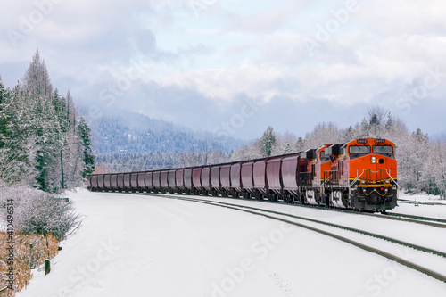 winter scene of locomotive pulling freight cars close to Whitefish, Montana with fresh snow in the foreground and surrounding foliage.