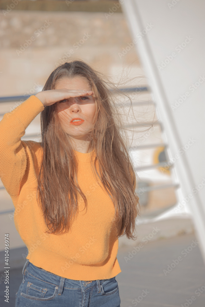 Beautiful girl in yellow jumper and blue trousers, blocking the sun from her face as she faces