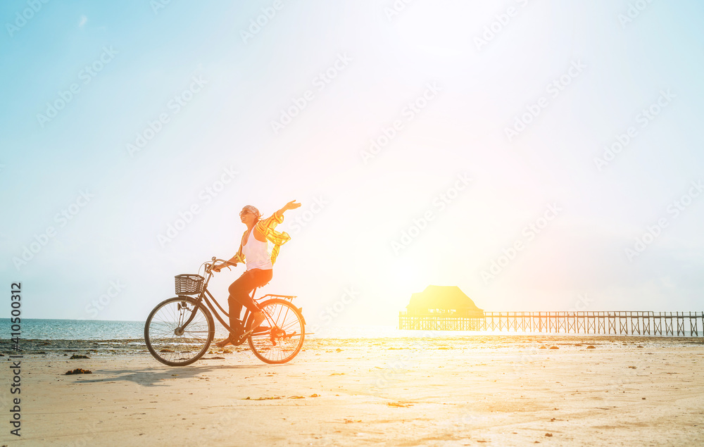 Obraz premium Young female dressed light summer clothes joyfully threw up her hand riding old vintage bicycle with front basket on the low tide ocean white sand coast on Kiwengwa beach on Zanzibar island, Tanzania.