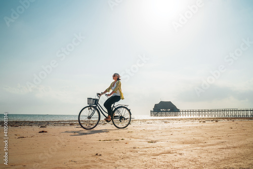 Young woman dressed light summer clothes riding old vintage bicycle with front basket on the lonely low tide ocean white sand coast on Kiwengwa beach on Zanzibar island, Tanzania.