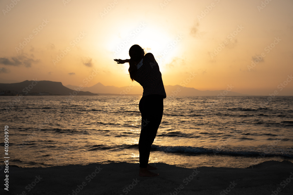 Silhouette of unrecognized young woman with protective surgical face mask performs yoga stretching exercises at the beach at dusk during covid-19 coronavirus pandemic