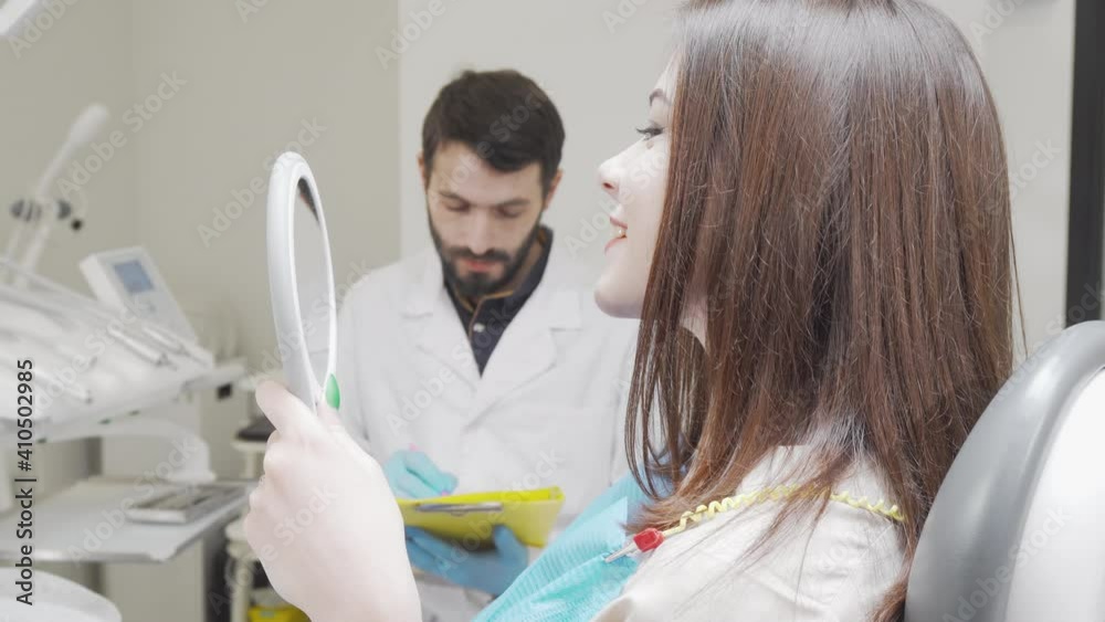Cheerful woman smiling to the camera after dental checkup. Young happy woman with healthy teeth looking in the mirror after dental treatment. Visiting dentist concept Cheerful woman smiling to the camera after dental checkup. Young happy woman with healthy teeth looking in the mirror after dental treatment. Visiting dentist concept