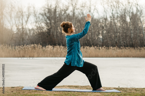 Young woman in comfortable, large clothes practicing yoga warrior I pose modified, in an urban park, by a lake. Concept: self care practices, movement, introspection, yoga outdoors
