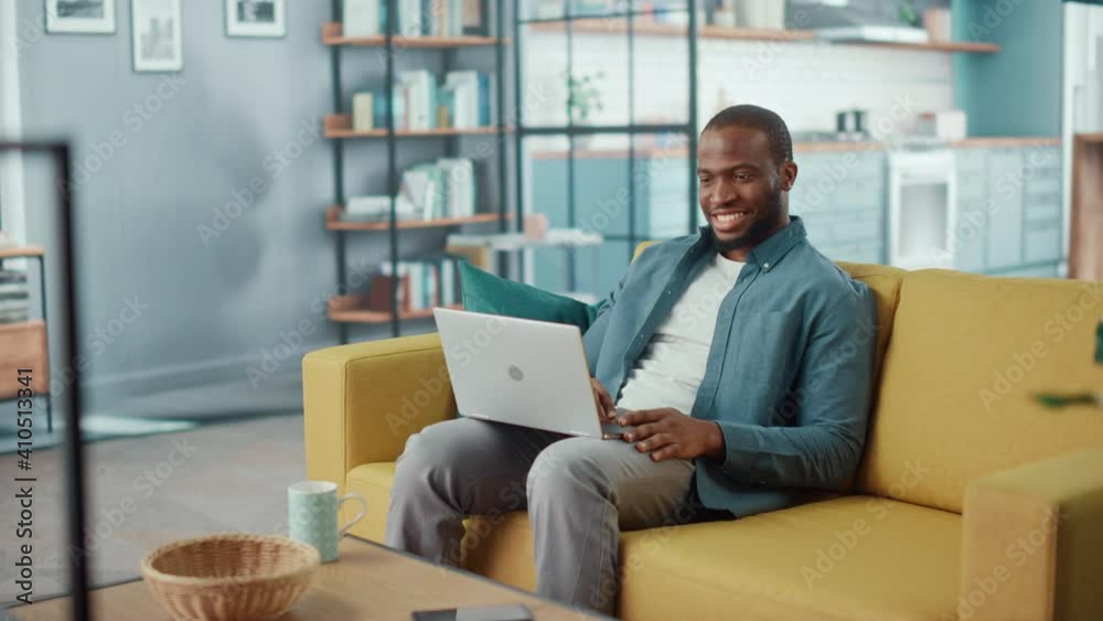 Handsome Black African American Man Working on Laptop Computer while Sitting on a Sofa in Cozy Living Room. Freelancer Working From Home. Browsing Internet, Using Social Networks, Having Fun in Flat. Handsome Black African American Man Working on Laptop Computer while Sitting on a Sofa in Cozy Living Room. Freelancer Working From Home. Browsing Internet, Using Social Networks, Having Fun in Flat.