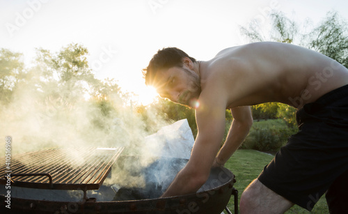 Barbecue in the garden at sunset. Portrait of a young caucasian adult grilling outdoors. The beautiful smoke and sensual shirtless man cooking. 