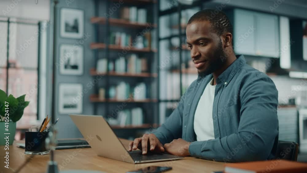 Handsome Black African American Man Working on Laptop Computer while Sitting Behind Desk in Cozy Living Room. Freelancer Working From Home. Browsing Internet, Using Social Network, Having Fun in Flat.