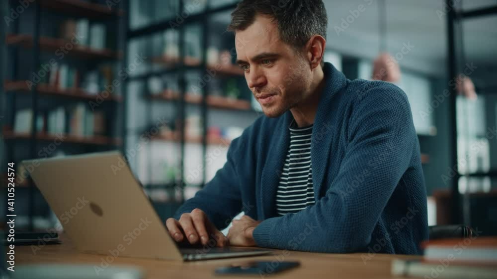 Handsome Caucasian Man Working on Laptop Computer while Sitting Behind Desk in Cozy Living Room. Freelancer Working From Home. Browsing Internet, Using Social Networks, Having Fun in Flat. Handsome Caucasian Man Working on Laptop Computer while Sitting Behind Desk in Cozy Living Room. Freelancer Working From Home. Browsing Internet, Using Social Networks, Having Fun in Flat.
