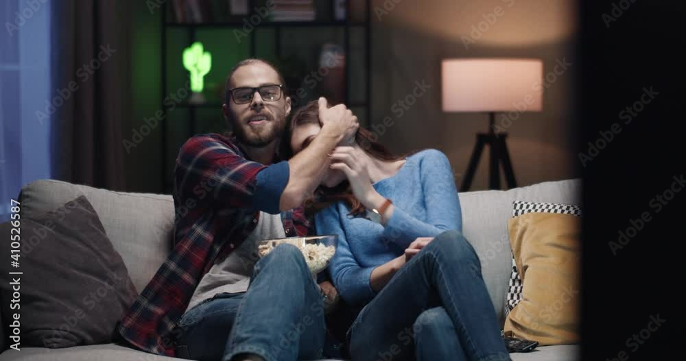 Young happy caucasian couple sitting on sofa at night at home and watching horror movie. Guy calms down scared girl. Young happy caucasian couple sitting on sofa at night at home and watching horror movie. Guy calms down scared girl.