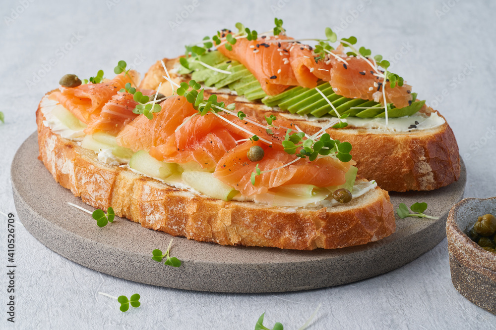 Two open sandwich, toast with salmon, cream cheese, avocado, cucumber slices on white concrete table. Morning healthy breakfast with fish and cup of drink, side view, close up Two open sandwich, toast with salmon, cream cheese, avocado, cucumber slices on white concrete table. Morning healthy breakfast with fish and cup of drink, side view, close up