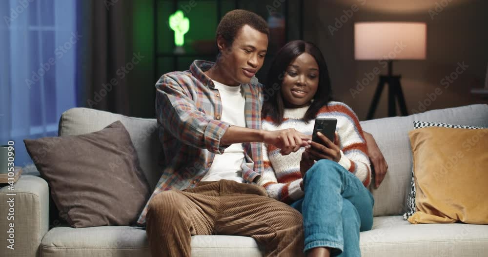 Young african american couple sitting at home on couch and flipping through the news feed on social networks using smartphone. Young african american couple sitting at home on couch and flipping through the news feed on social networks using smartphone.