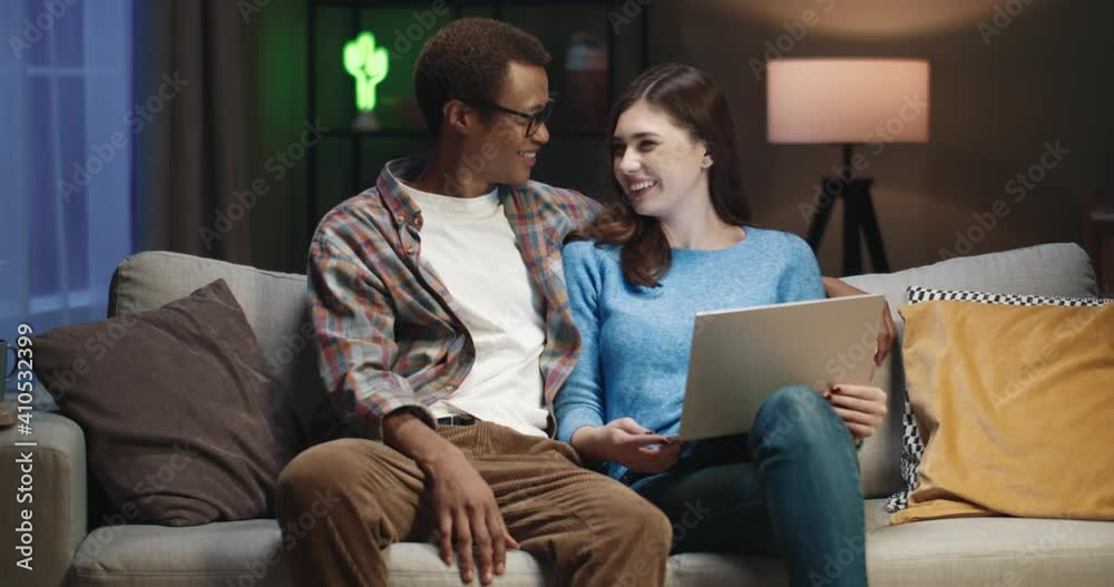 Young happy multinational couple sitting at home in evening on couch and spending free time watching interesting news on laptop. Young happy multinational couple sitting at home in evening on couch and spending free time watching interesting news on laptop.